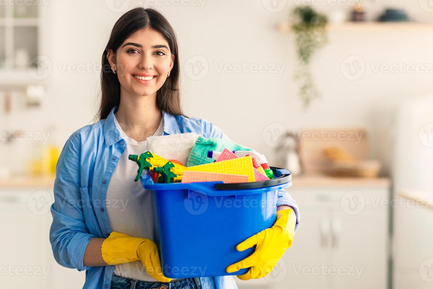 Housekeeping Concept. Portrait of smiling woman cleaner holding bucket with detergents and supplies, posing and looking at camera standing in kitchen, wearing rubber gloves, blurred background photo