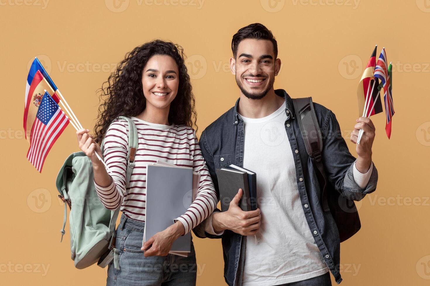 Education Abroad. Happy Young Arab Man And Woman Holding Stack Of International Flags, Smiling Middle Eastern Students With Backpacks And Workbooks Posing Over Yellow Studio Background, Copy Space photo
