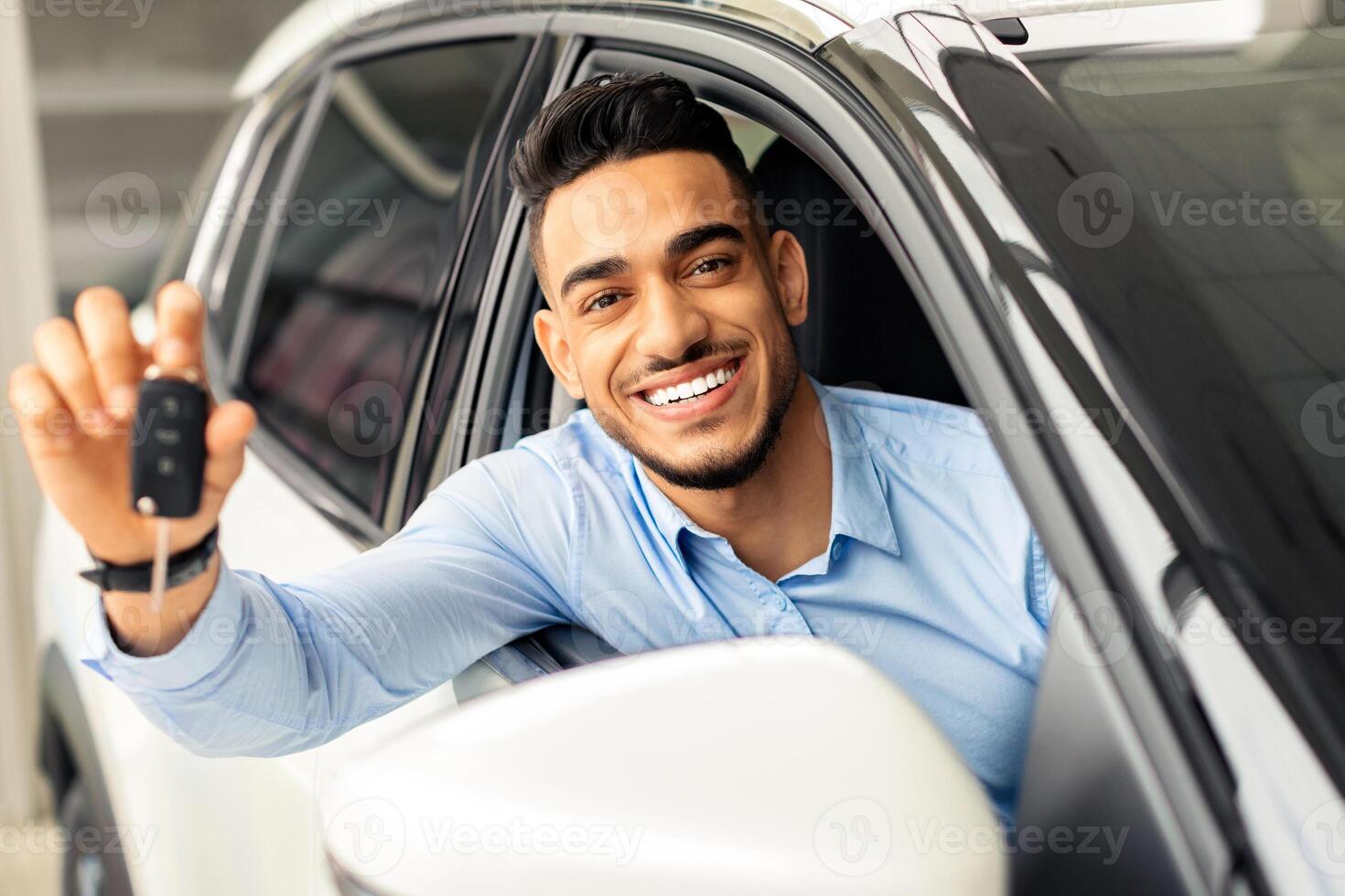 Cheerful arab man holding key from auto, buying brand new car in showroom. Happy middle-eastern guy buying new automobile, sitting inside black car and showing automatic key at camera, smiling photo