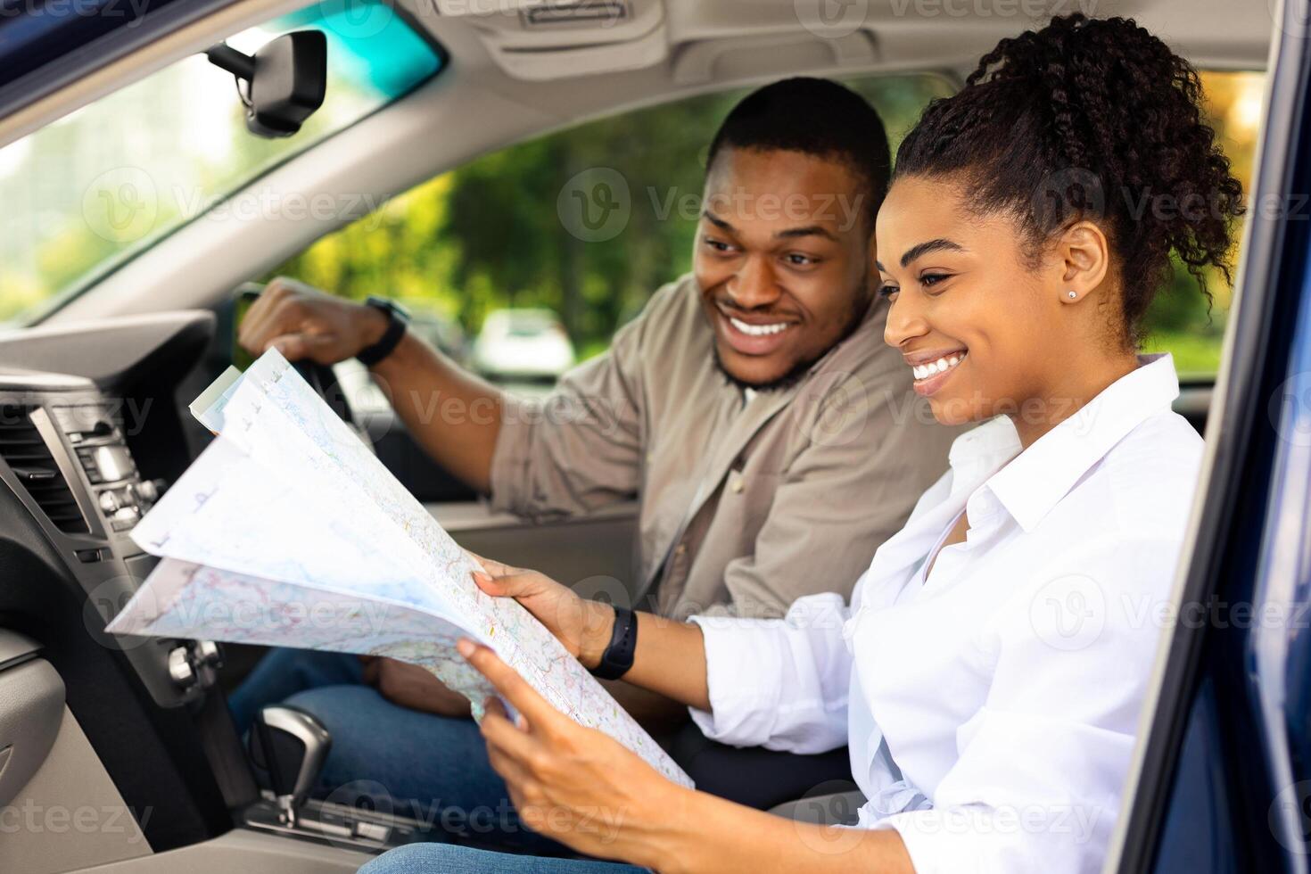 Happy Black Couple Sitting In Car Holding Road Map Choosing Travel Destination For Summer Travel Together. Auto Tourism And Navigation Concept. Selective Focus, Side View photo