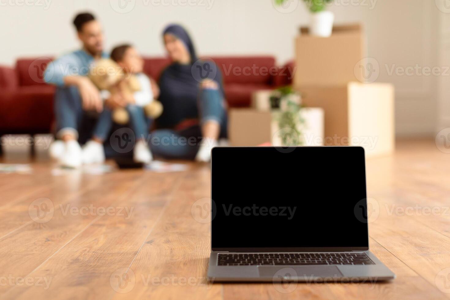 Renovation And Technology Concept. Selective focus on laptop with black empty screen for mock up, free copy space. Smiling dad, mom and daughter sitting on floor in living room in blurred background photo