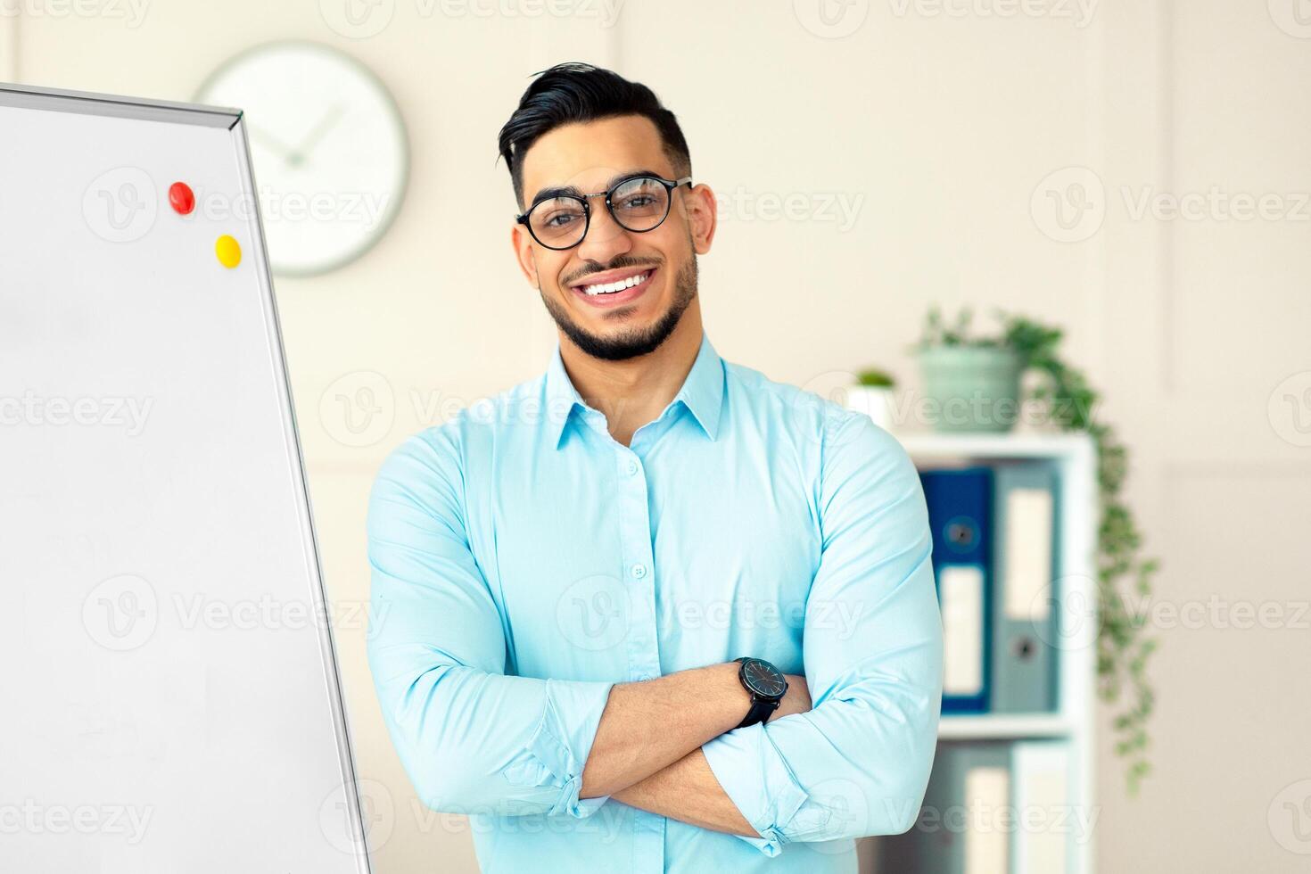 Distance learning. Happy Arab guy teacher giving online English class, standing with crossed arms near blackboard, smiling at camera in home office. College tutor explaining grammar rules to students photo
