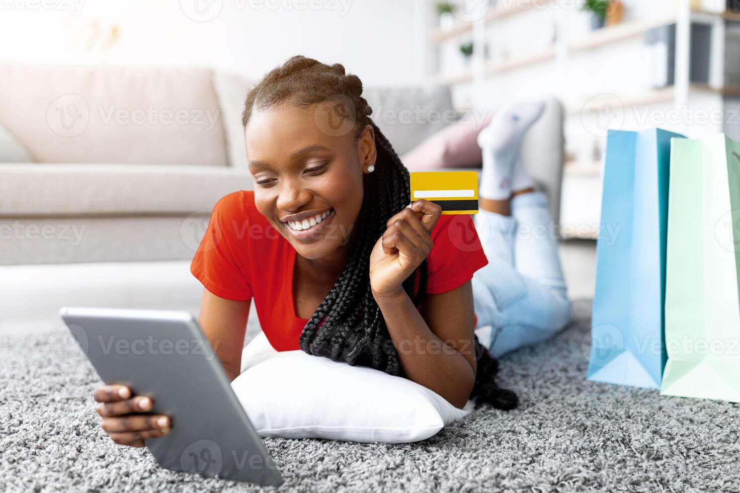 Cheerful black lady lying on floor with tablet pc and credit card, shopping on web from home. Smiling young woman with gift bags purchasing stuff online in web store. Sales, discounts, e-commerce photo