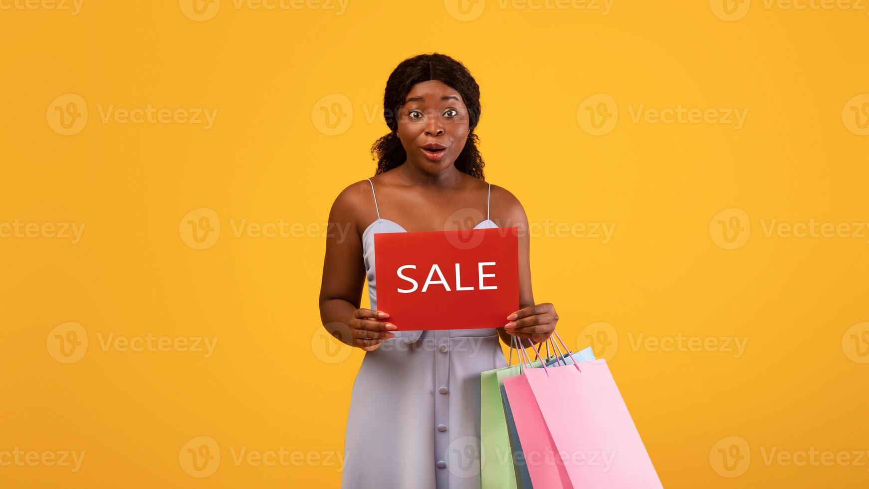 Surprised millennial black woman holding shopping bags and SALE sign over orange studio background. Young African American female promoting seasonal discounts, feeling excited photo