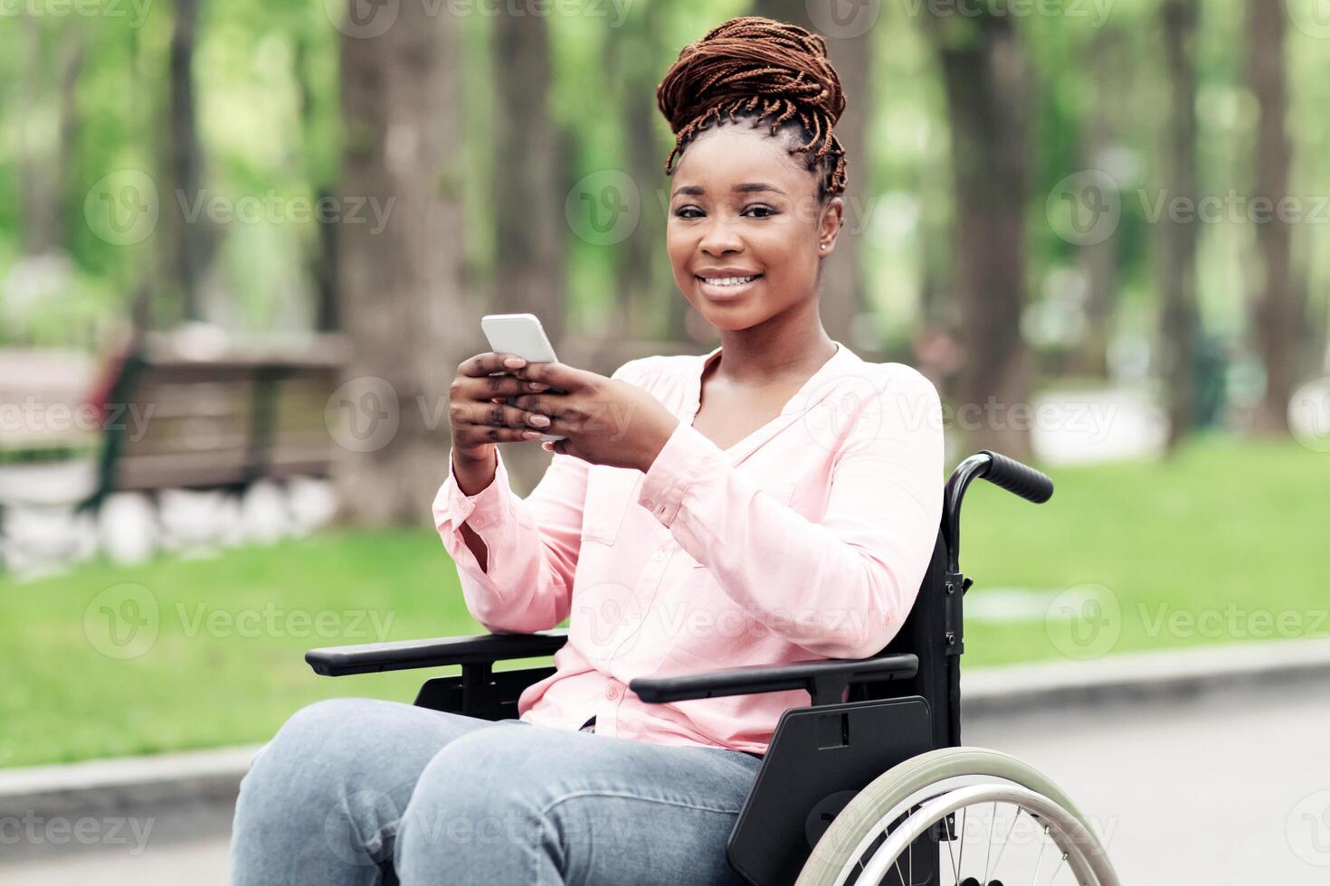 Cheery young black woman in wheelchair using mobile phone, checking messages at spring park, copy space. Millennial African American lady with cellphone posting in social network, browsing web photo