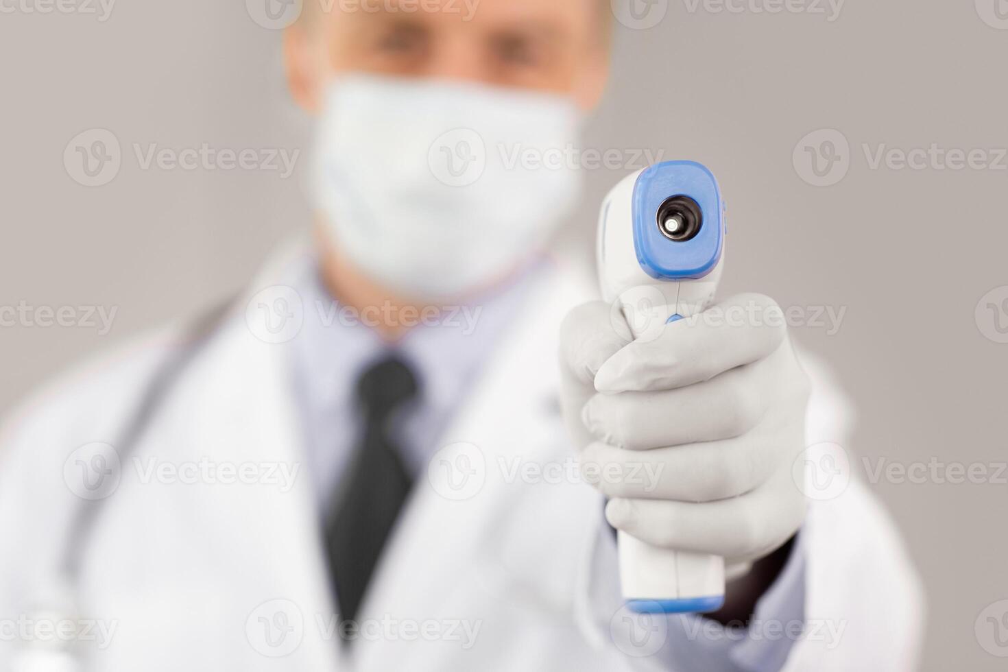 Concept of Covid-19 Prevention. Man in medical mask measuring body temperature. Doctor holding digital infrared non-contact thermometer in hand, pointing at camera. Coronavirus Symptoms photo