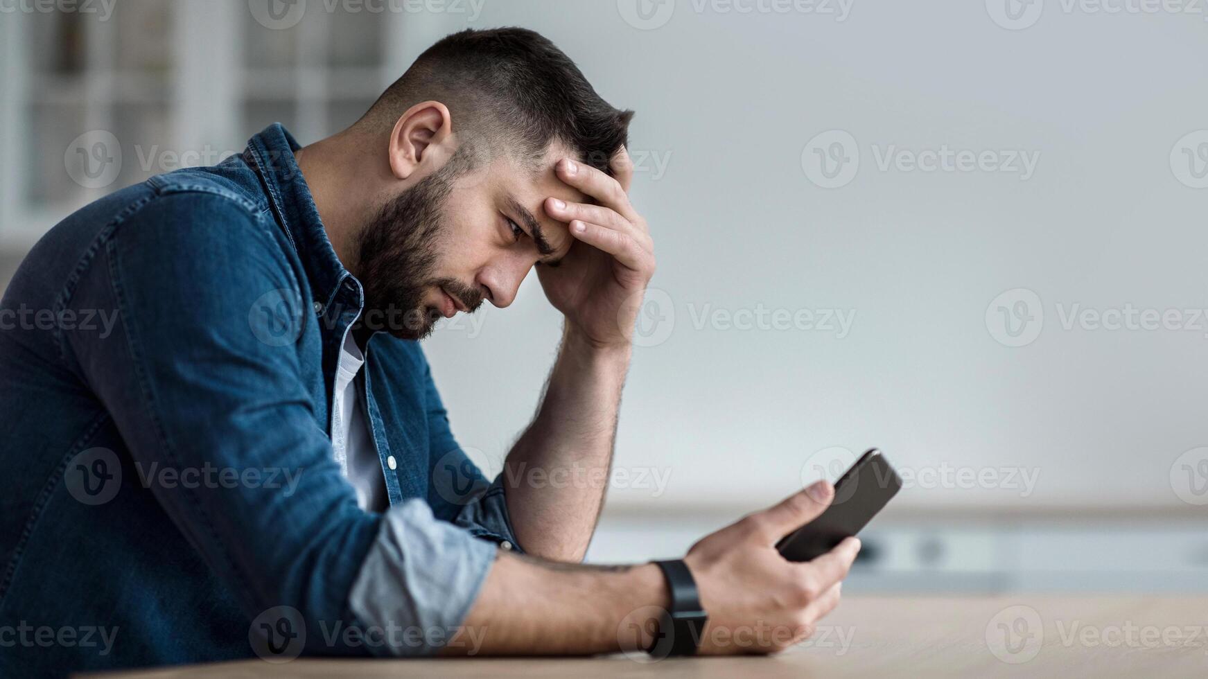Waiting for call, problems and difficulties, stress and no answer. Unhappy sad disappointed young bearded guy looks at smartphone and presses hand to head, sits at table in kitchen interior, profile photo