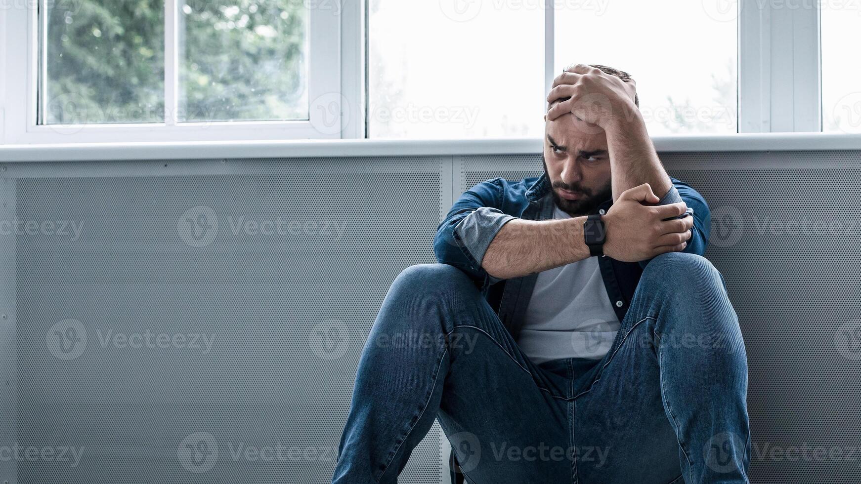 Man feeling depressed and desperate, crying alone at home, suffering, emotional pain, unhappy, mental health issues. Sad young bearded guy sits on floor near large window and holds his head with hand photo