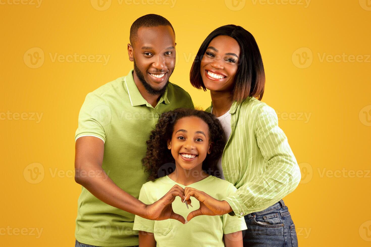 Happy loving family. Black parents holding hands in heart shape sign, posing with liitle daughter over yellow background and smiling together to camera photo