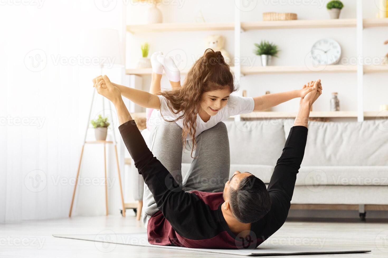 Adorable little curly girl exercising with her daddy at home, middle-eastern young man father making airplane with his cute daughter, laying on fitness mat in living room, lifting kid up, copy space photo