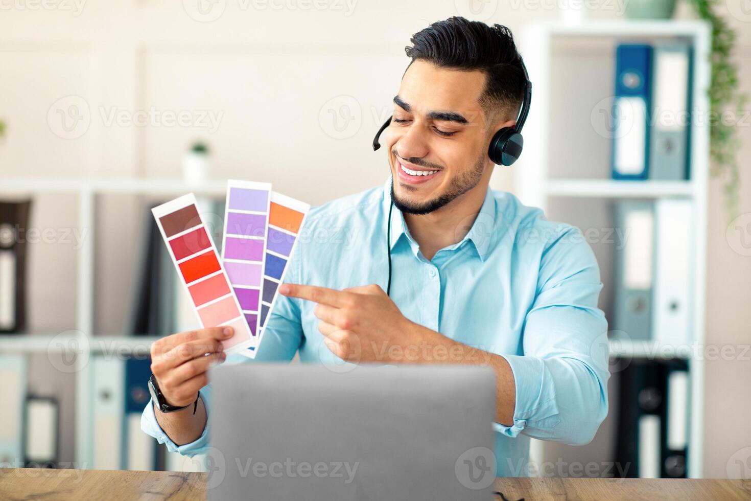 Millennial Arab guy showing color swatches at laptop webcam in office, presenting graphic design project during remote business meeting. Young guy designer choosing gamma for future house interior photo