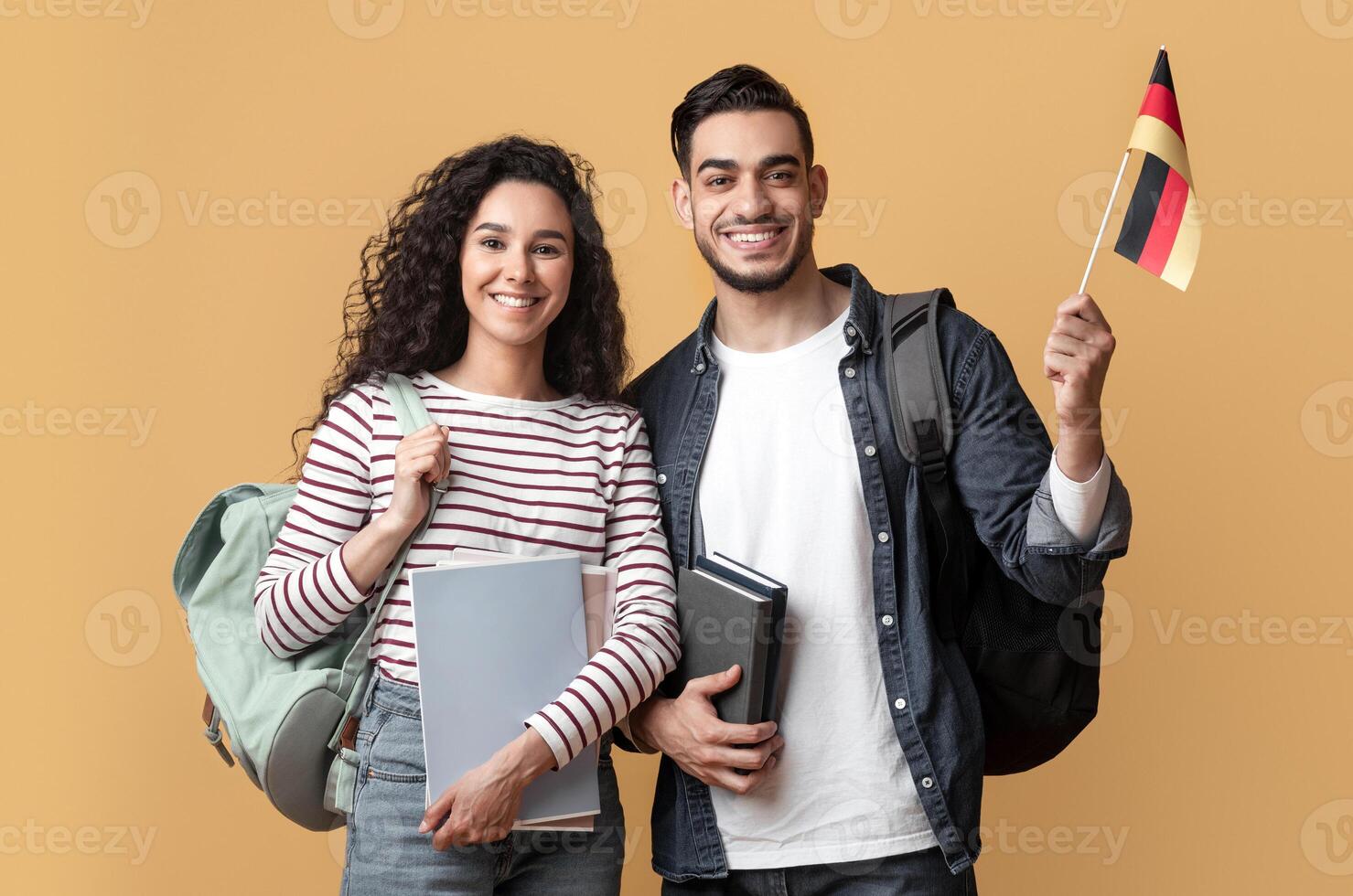 International Education. Middle Eastern Student Couple With Backpacks Holding German Flag, Portrait Of Smiling Arab Man And Woman With College Workbooks Posing On Yellow Studio Background, Copy Space photo