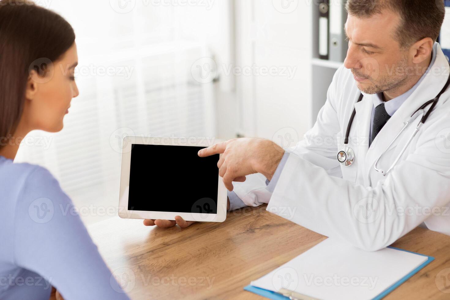 Medical Test And Examination Results. General practitioner showing patient digital tablet with black blank screen, discussing treatment, sitting at the table at office, pointing at monitor photo