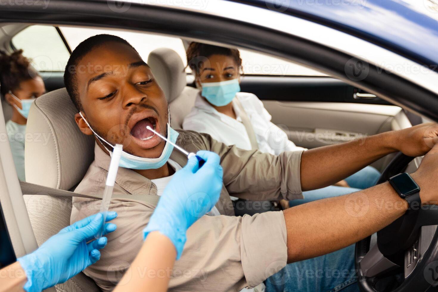 Black Male Driver Getting Tested For Covid-19 Sitting In Automobile. Medical Worker Testing Family Of Three In Car, Wearing Protective Gloves. Healthcare And Transportation. Selective Focus photo