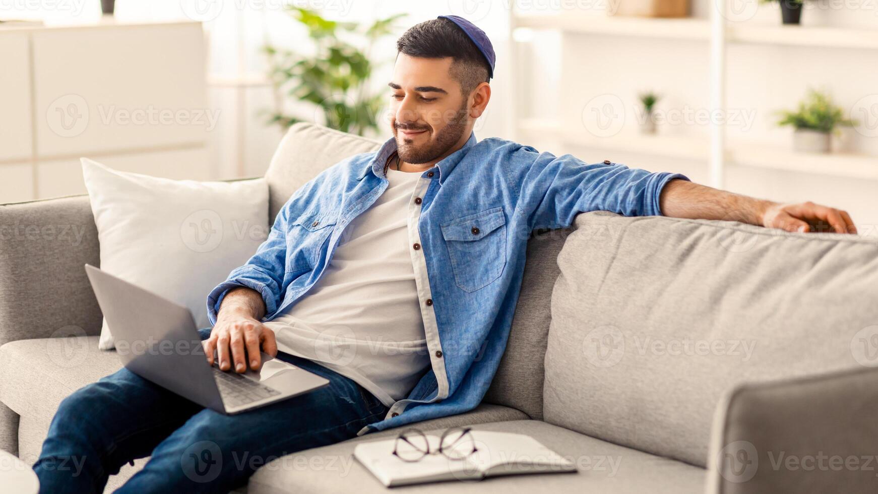 persona de libre dedicación jornada laboral. sonriente milenario hombre empleado estudiante trabajando estudiando desde hogar sentado en sofá en vivo habitación. masculino utilizando ordenador portátil comprometido en electrónico papeleo haciendo distante tarea foto