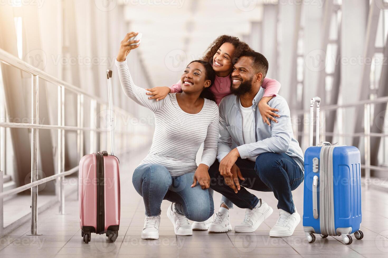 Selfie At Airport. Happy Young Black Family Of Three People Taking Self Picture With Mobile Phone In Airport Terminal, Posing At Camera, Traveling Together. Daughter Embracing Parents From Behind photo
