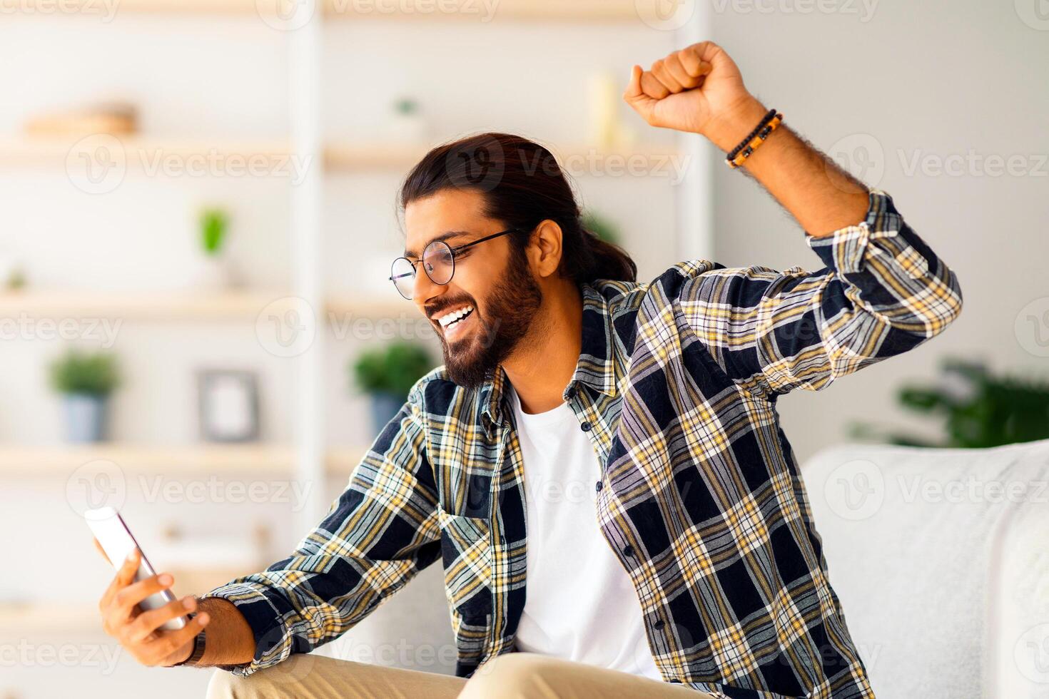Emotional long-haired indian guy with glasses sitting on couch, using smartphone, raising hand up, happy millennial arab man chilling at home, gambling or trading, side view, copy space photo
