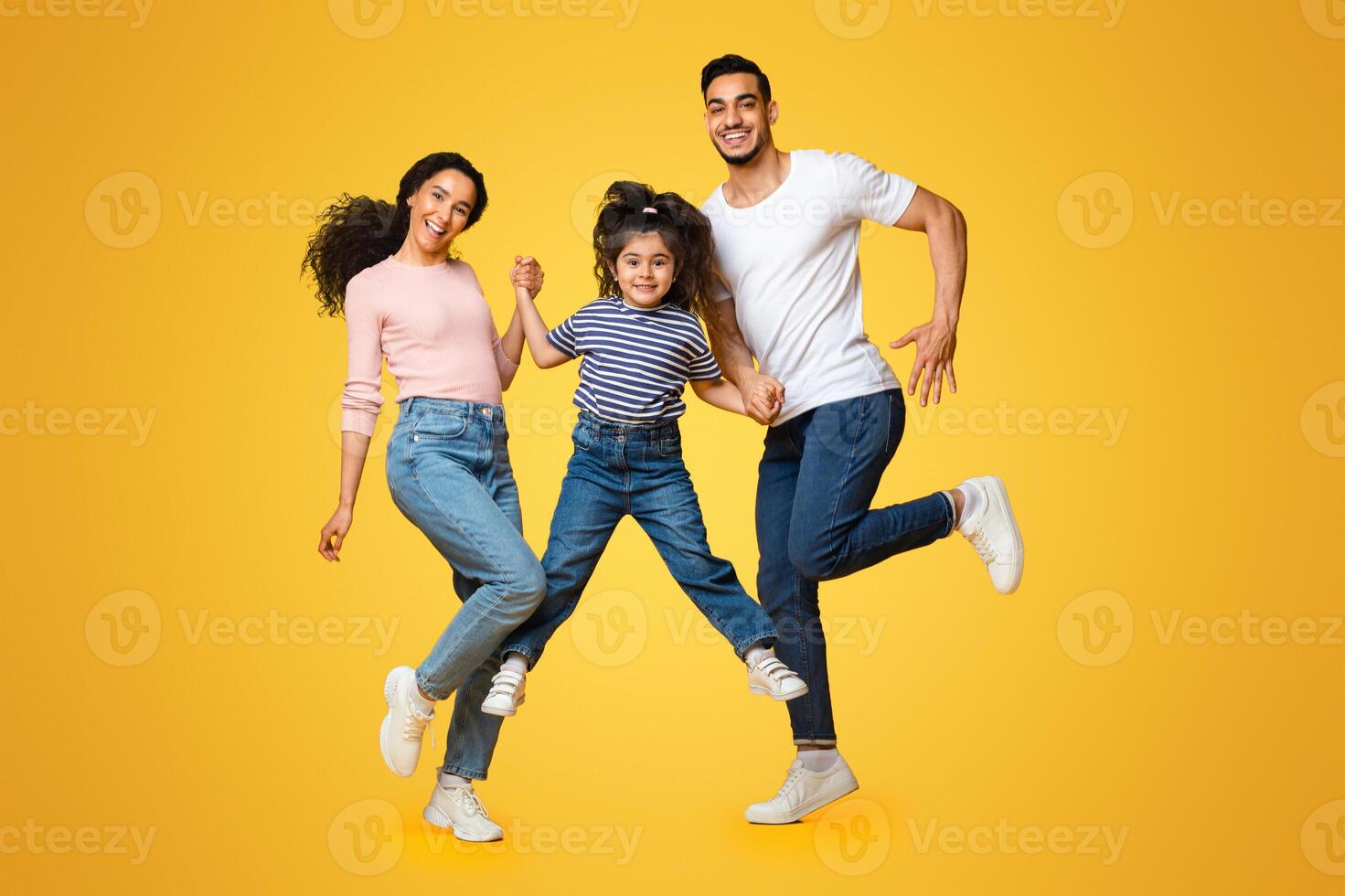 Portrait Of Cheerful Excited Middle Eastern Family Of Three Holding Hands While Jumping In Air Over Yellow Background In Studio, Happy Arab Parents And Little Daughter Having Fun Together, Copy Space photo