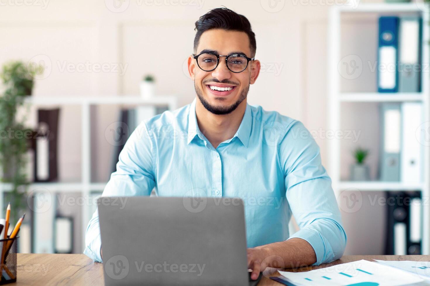 Happy young Arab businessman in glasses sitting at desk, working on laptop at company office. Successful smiling CEO having online job, typing on pc keyboard, having call photo