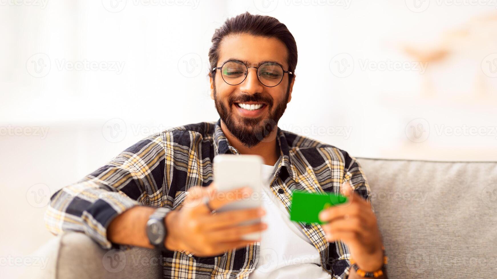 Cheerful young arab man with credit card and cellphone sitting on couch at home, closeup, copy space. Happy indian guy freelancer shopping online, using smartphone and holding card, e-commerce photo