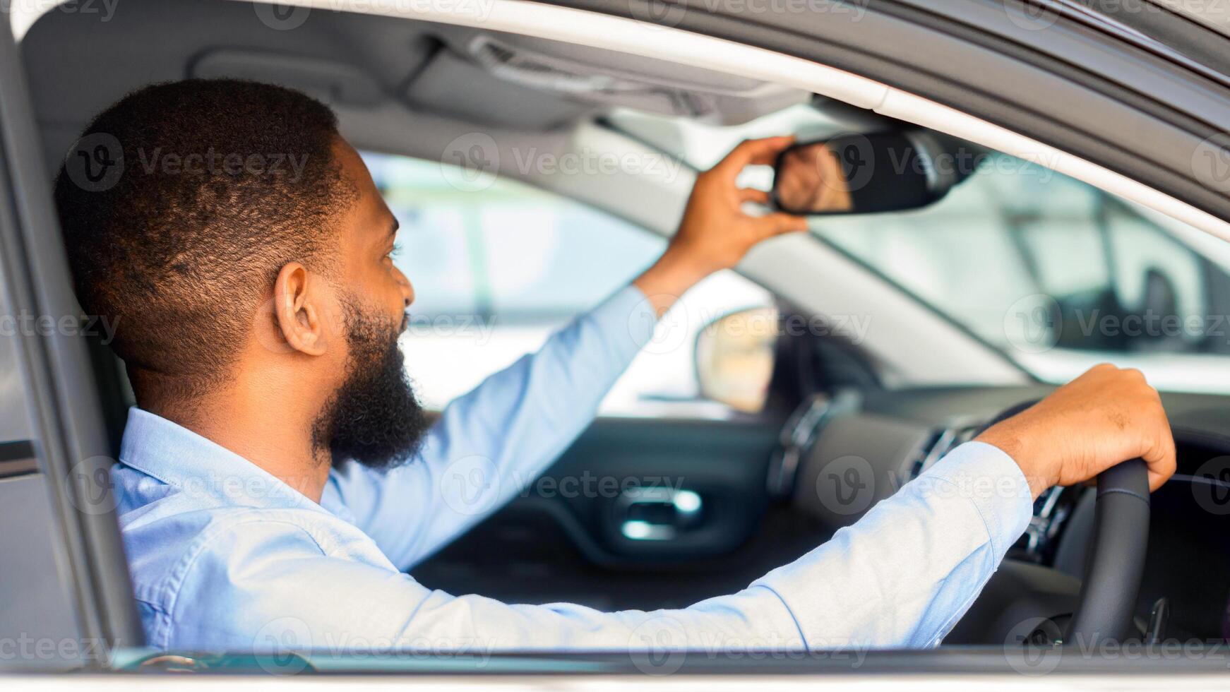 Young Black Man Choosing New Vehicle, Sitting Inside Of Car And Adjusting Rear View Mirror, Happy African American Male Customer Purchasing Automobile In Modern Dealership Center, Closeup Shot photo