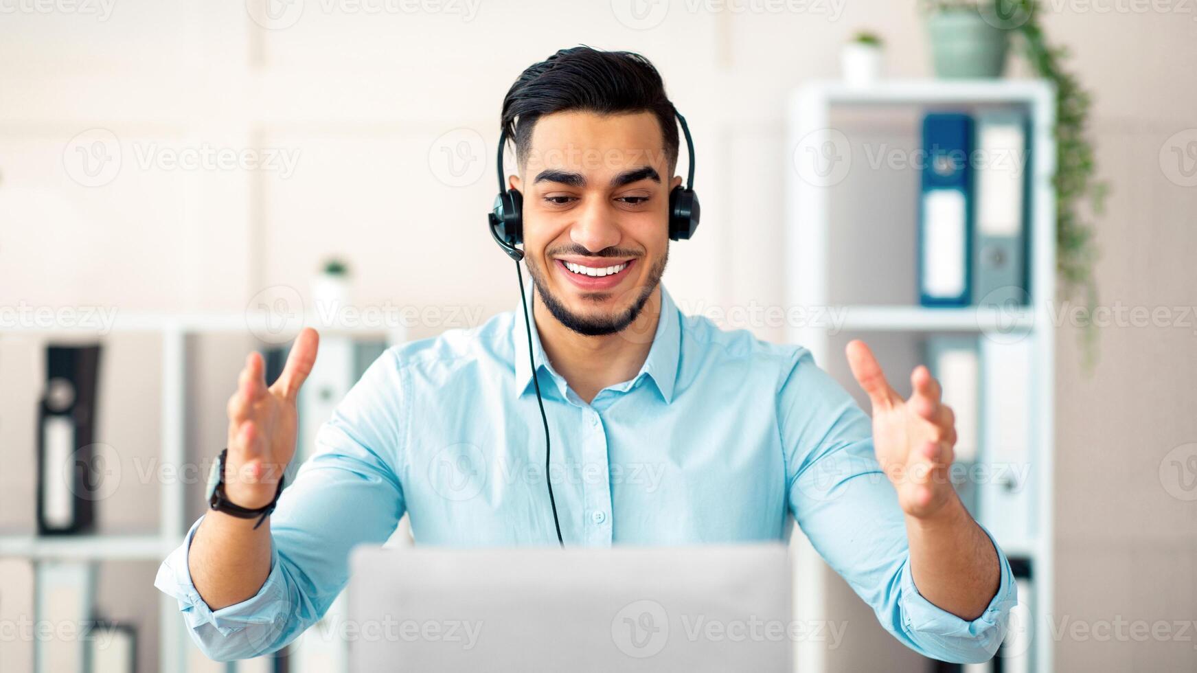 Telecommuting concept. Happy Arab man with headphones working online on laptop, having call at office. Young guy using computer, communicating on webcam, having business meeting photo