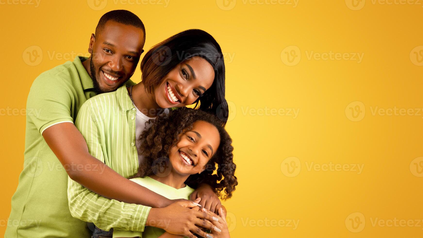 Portrait of happy black family of three with cute daughter, joyful african american parents and their female child embracing and smiling together to camera, posing over yellow background photo