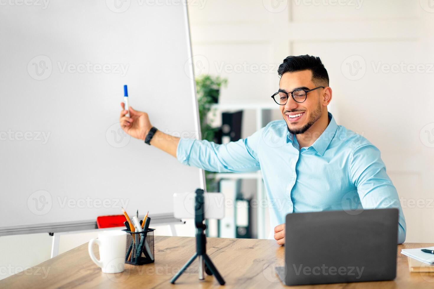 Cyberlearning, remote education concept. Happy Arab guy teacher giving online English lesson, using smartphone and laptop, pointing at blackboard with grammar rules at home office photo