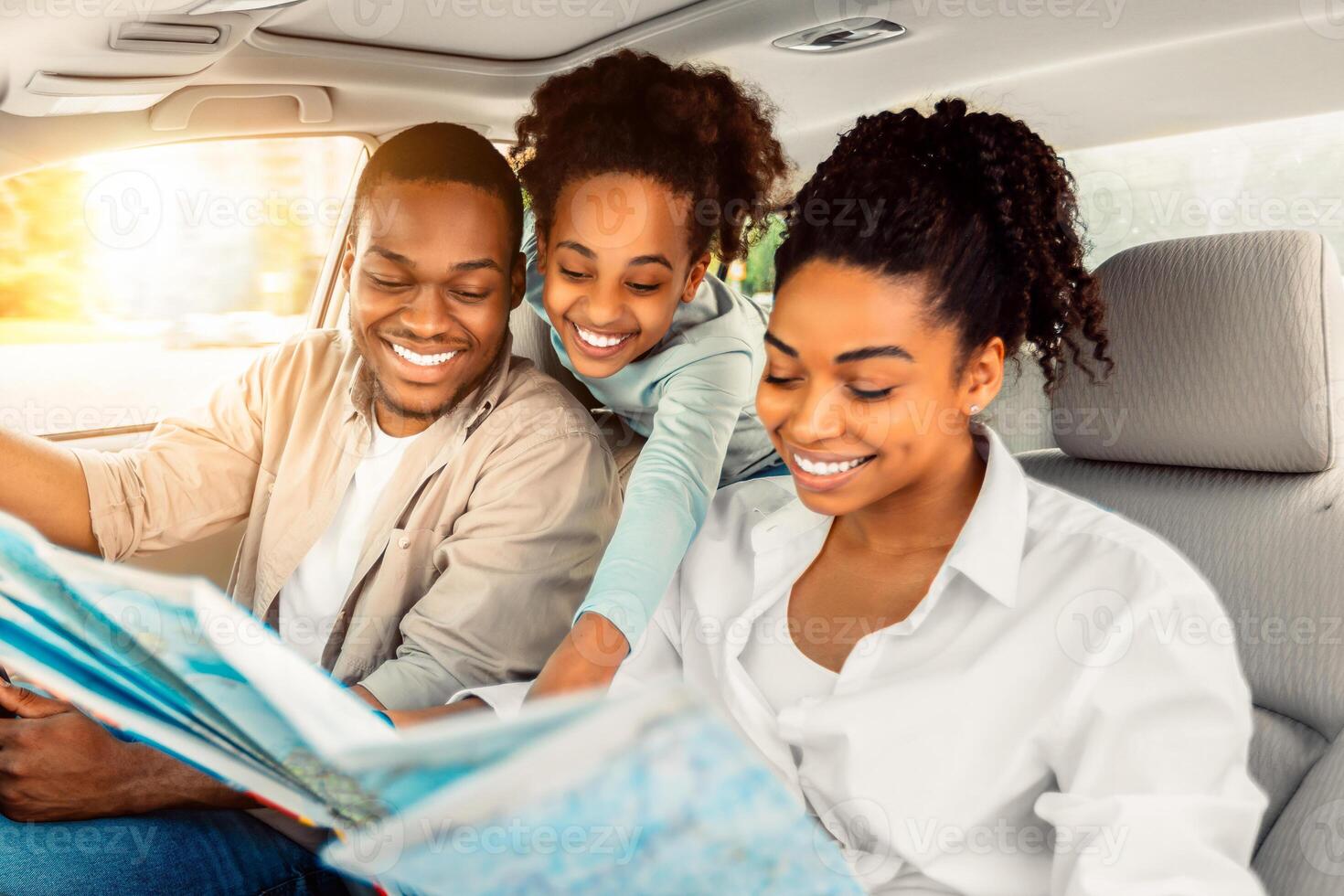 Cheerful African American Family Looking At Road Map Sitting In Car. Parents And Daughter Choosing Destination For Summer Road Trip Together. Local Tourism Concept. Selective Focus photo