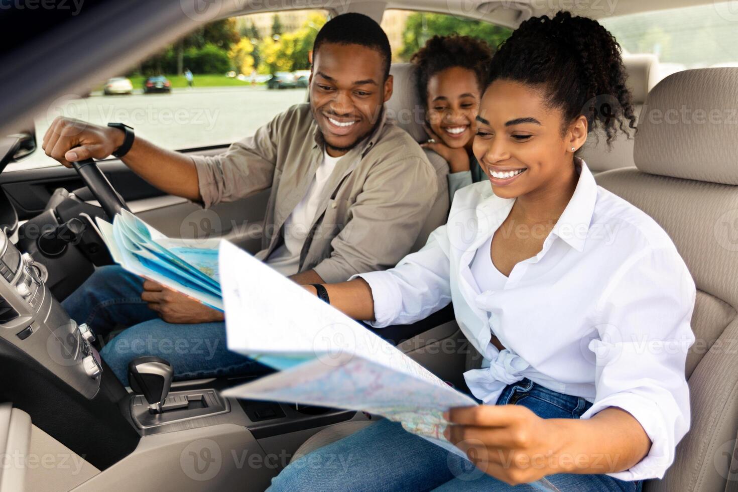 Happy Black Family Sitting In Car Looking At Road Map Planning Summer Trip. African American Parents And Daughter Choosing Destination Traveling Together. Auto Tourism Concept. Selective Focus photo