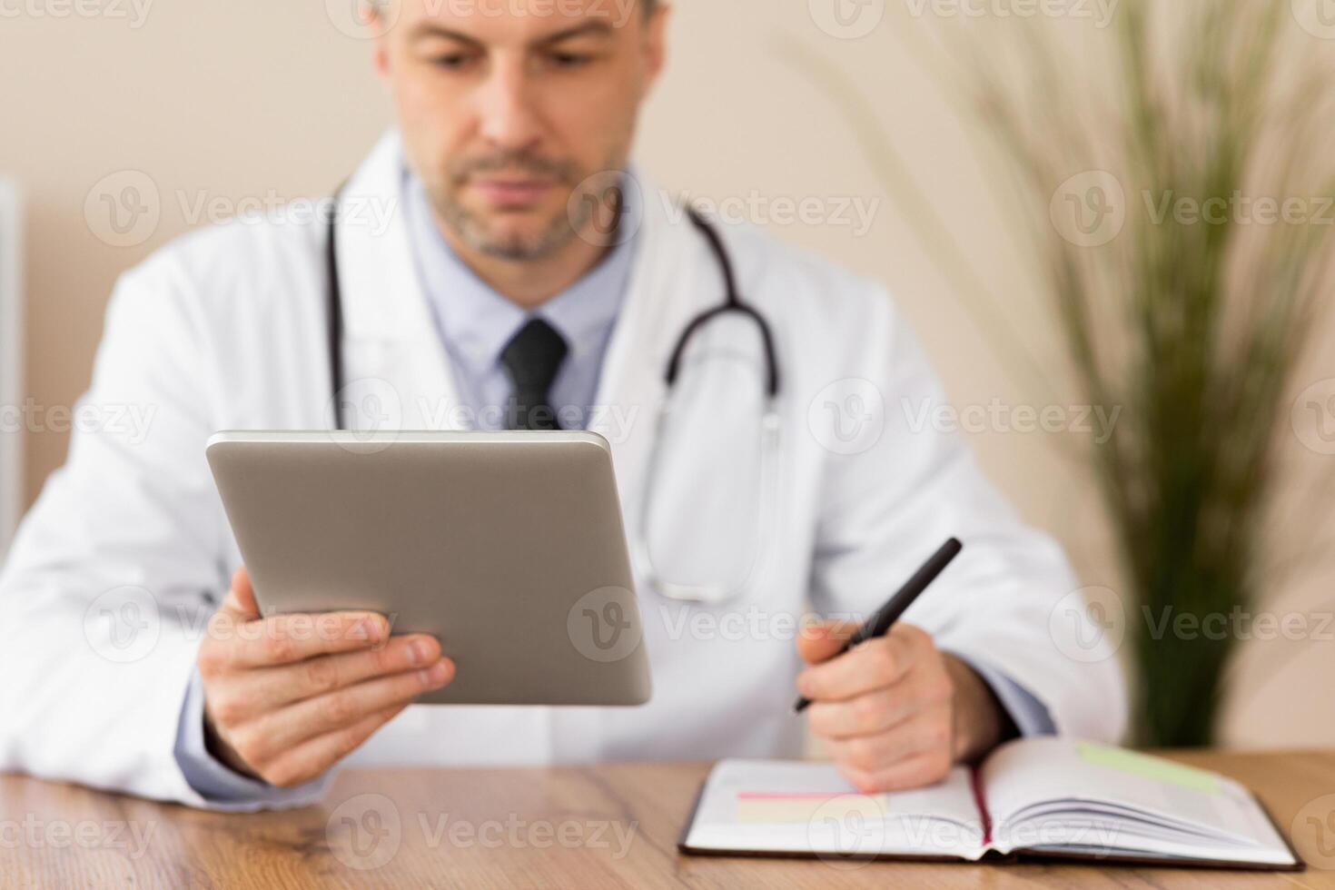 Close up of medical practitioner filling in paperwork sitting at desk in his in office and holding tablet photo