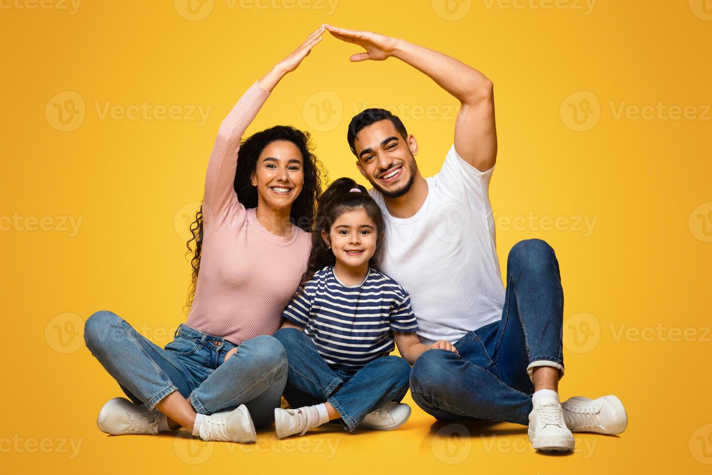 Family Insurance Concept. Happy arab parents making symbolic roof of hands above little daughter, middle eastern mom and dad sitting together with child on floor over yellow background in studio photo