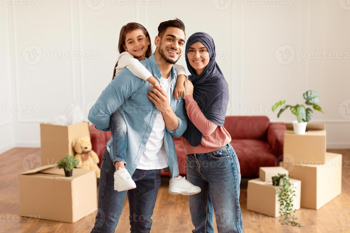 Celebrating Moving Day. Portrait of happy middle eastern dad, mom in headscarf and daughter posing in new apartment, standing in empty living room among cardboard boxes, father riding girl on back photo