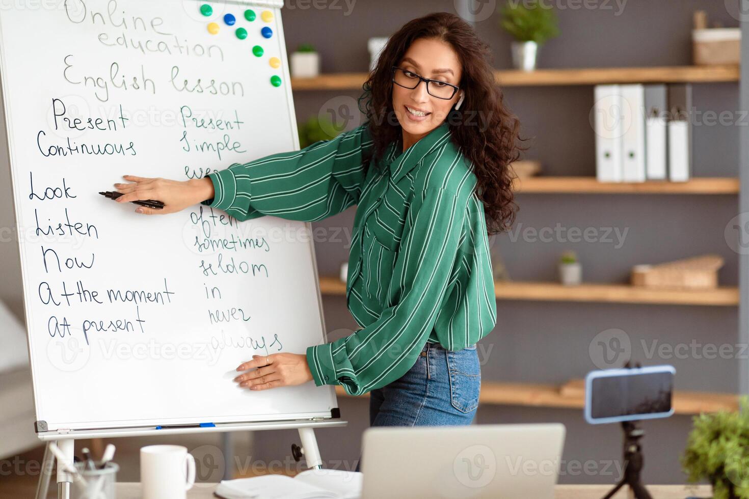 Online Learning Concept. Portrait of young lady teacher pointing at whiteboard, explaining grammar rules and tenses. Tutor recording lesson using mobile phone on tripod, live streaming course photo