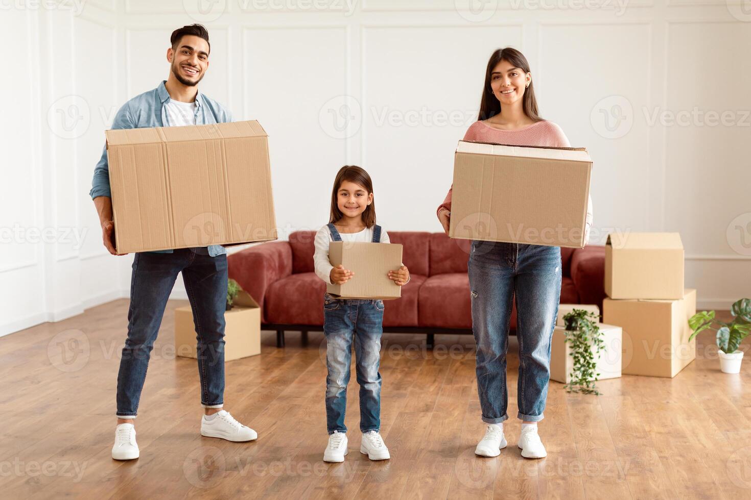 Happy young family of three moving to new apartment, standing together in living room, holding big carton paper boxes full of their belongings, looking at camera, posing and smiling, full body length photo