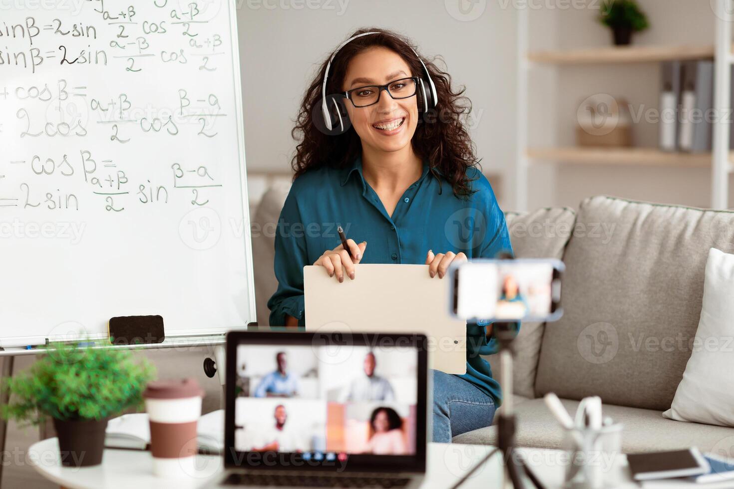 Studying Online. Smiling lady teacher in eyeglasses and headphones giving lesson on web, using smartphone and pc. Millennial lecturer having call with students recording vlog photo