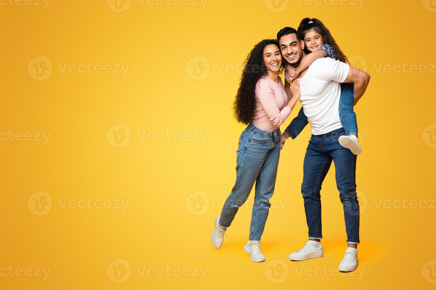 Family Time. Portrait Of Cheerful Middle Eastern Parents And Little Daughter Posing Together Over Yellow Studio Background, Happy Arab Mom, Dad And Cute Female Child Embracing And Smiling At Camera photo