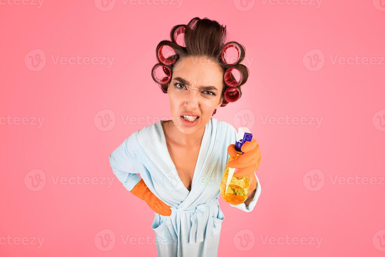 Angry Housewife With Curlers Holding Detergent Bottle Looking At Camera Standing Over Yellow Studio Background. High-Angle Shot photo