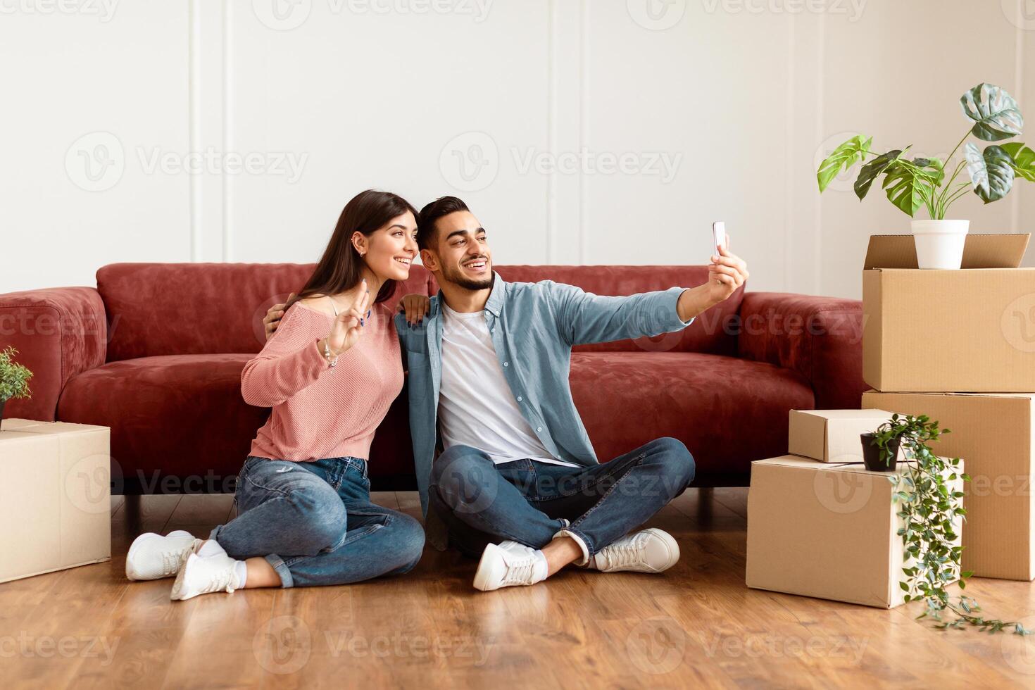 Happy young couple taking selfie in their new apartment, celebrating moving day, sitting on the floor near carboard boxes and plants. Smiling woman making showing v victory peace sign gesture photo