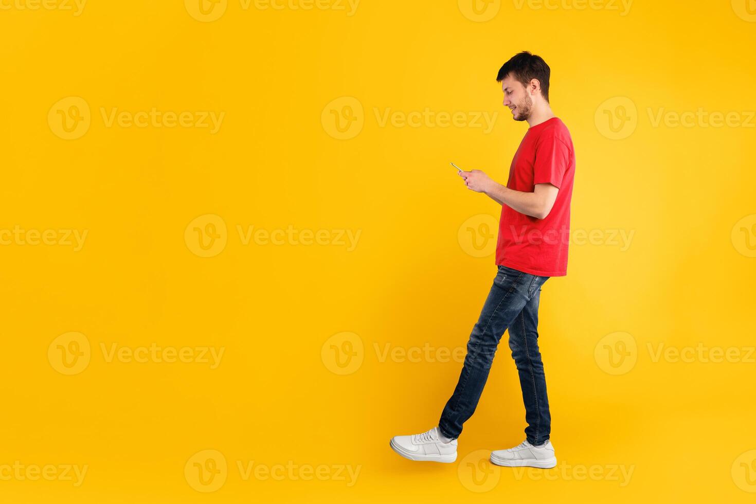 Communication, Technology And Lifestyle Concept. Full length portrait of handsome smiling young man using mobile phone, walking isolated over blue studio background, vertical shot photo