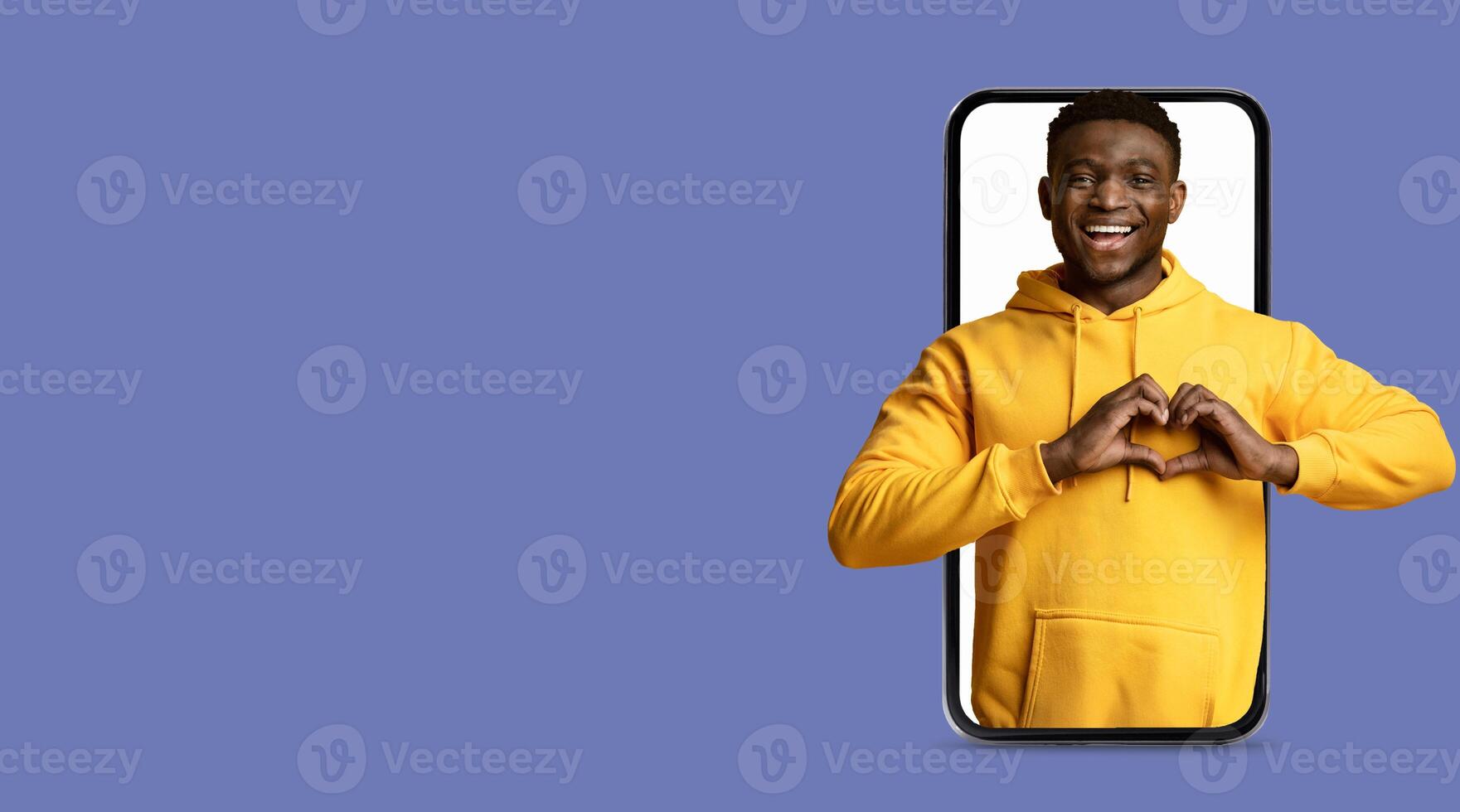 A young man wearing a yellow hoodie smiles as he creates a heart shape with his hands while interacting with a large smartphone screen. The background is a solid color, enhancing the focus on him. photo