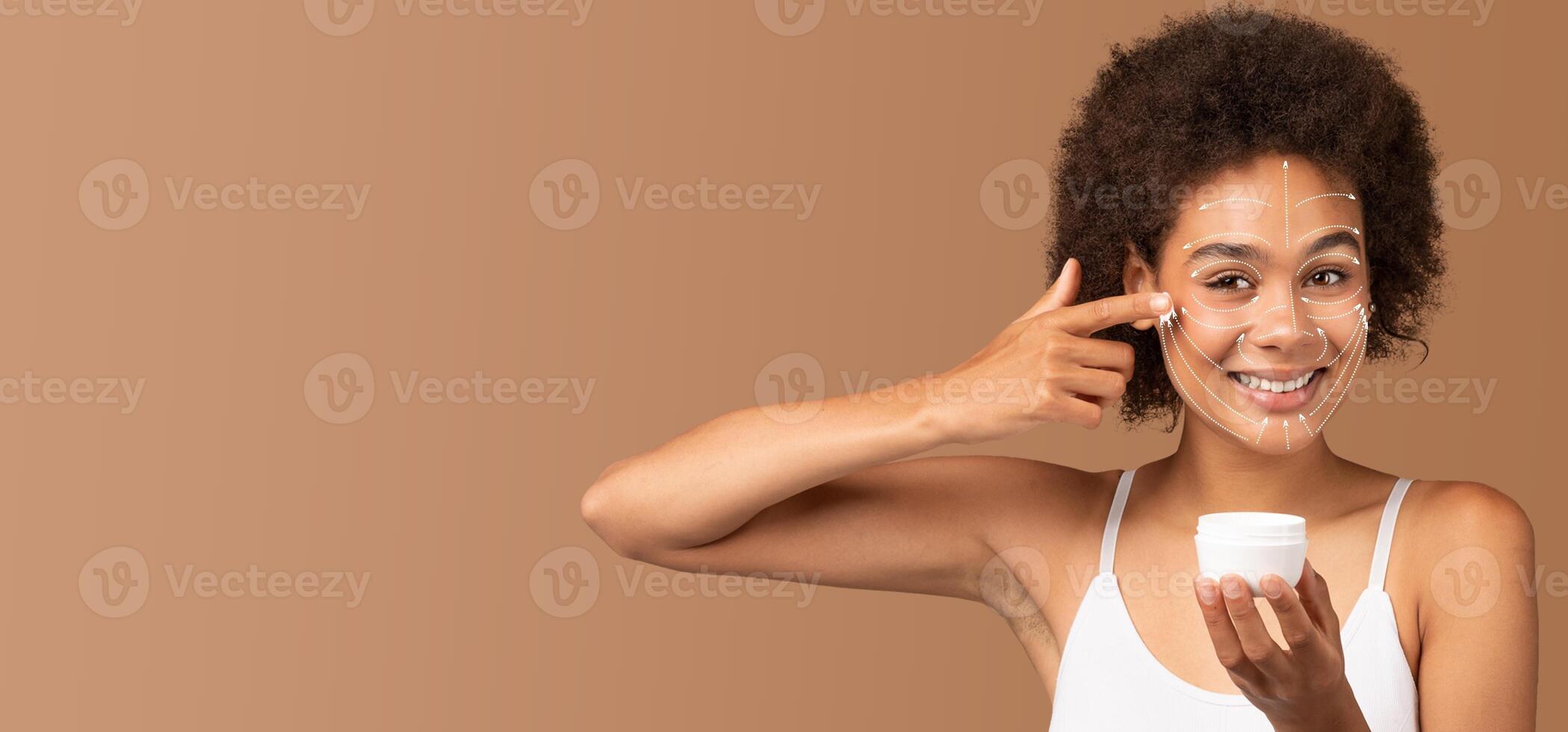 African American woman is happily applying skincare cream on her face. She is smiling, showcasing her beauty routine in a simple, well-lit environment. The focus is on pampering and self-care. photo
