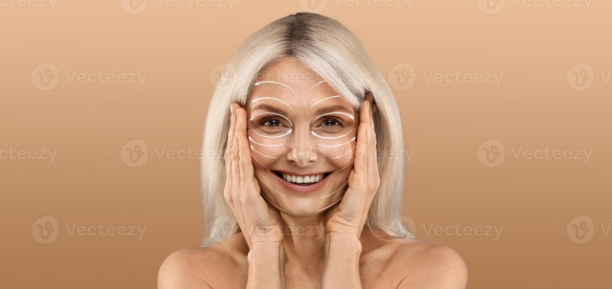 A smiling woman engages in her skin care routine, gently applying treatment around her face while standing against a warm, neutral background. The mood is relaxed and pampering. photo