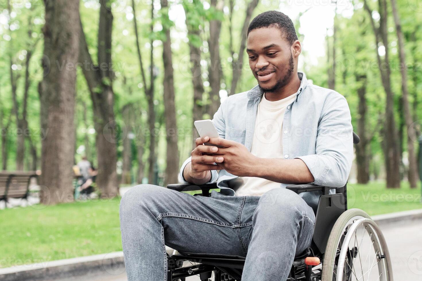 Full length of young black man in wheelchair using mobile phone, checking messages at park, free space. Millennial African American guy with cellphone browsing social network, surfing web photo