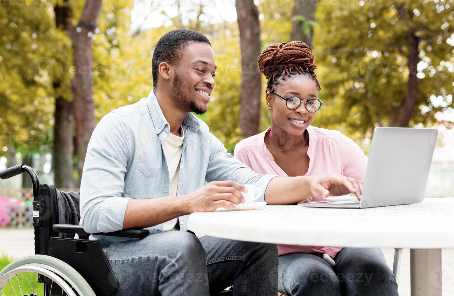 Cheerful impaired black man with female colleague using laptop together in outdoor cafe at autumn park, working on online business project, attending meeting remotely photo