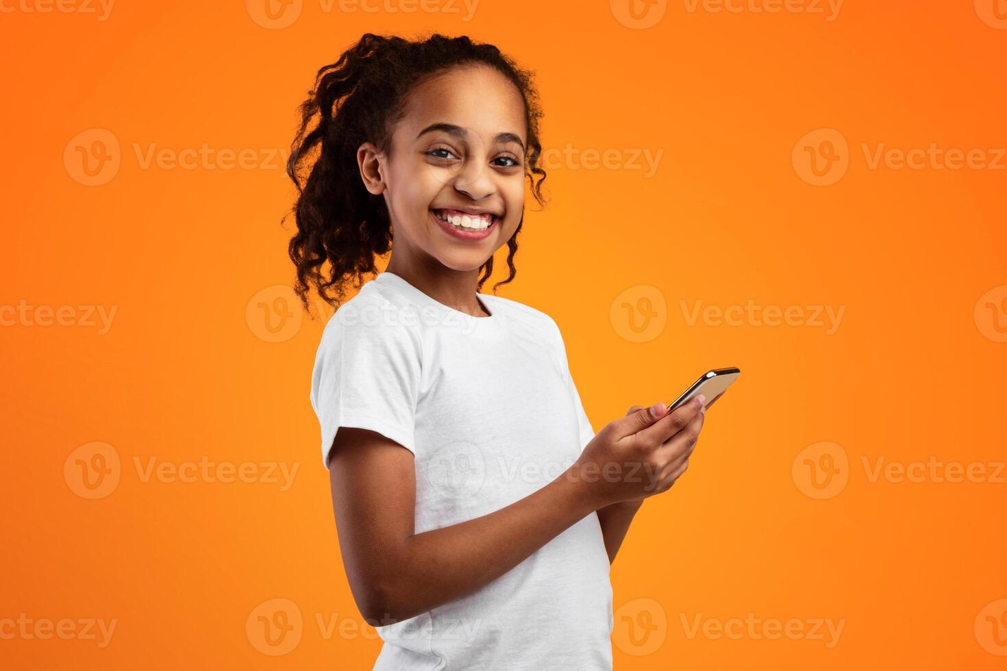 Portrait of smiling African American girl holding smartphone and looking at camera, standing isolated on yellow studio background. Happy black kid chatting online, typing sms, using app, banner photo