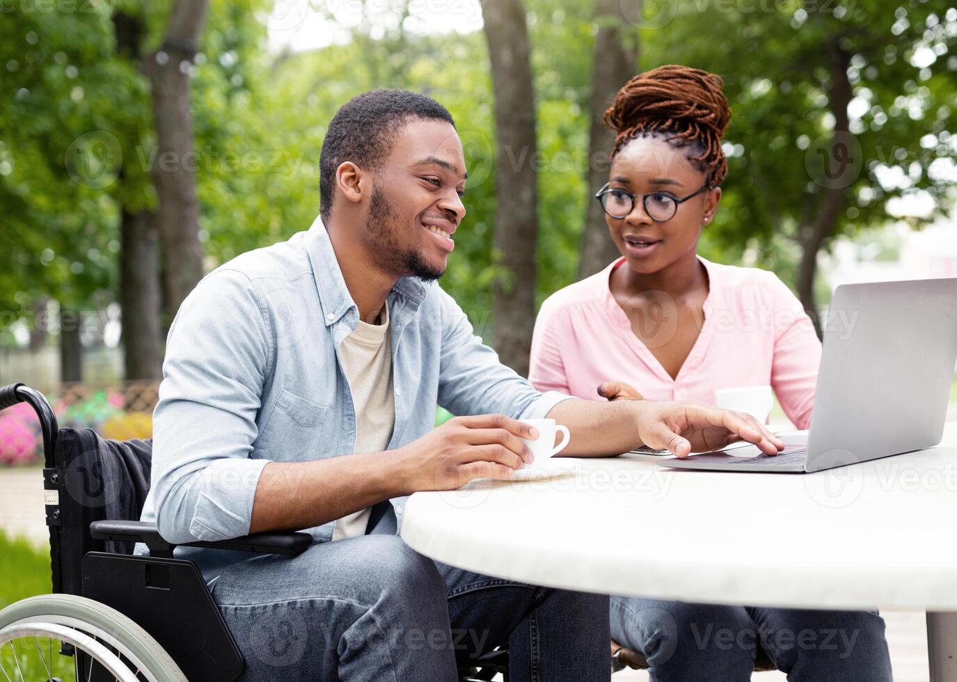 Happy disabled black man and his female coworker using laptop together at table in outdoor cafe, having coffee while working on online business project at park, copy space photo