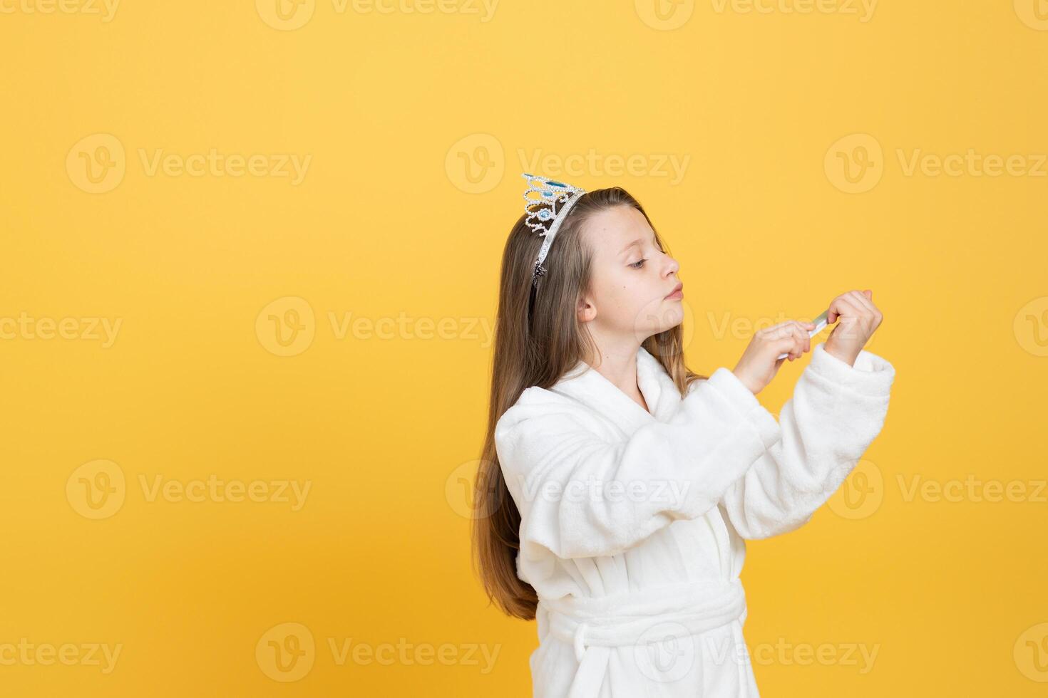 A preteen girl dressed in a cozy white robe doing manicure while wearing a stylish tiara. The vibrant yellow background creates a cheerful atmosphere, highlighting her playful spirit. photo