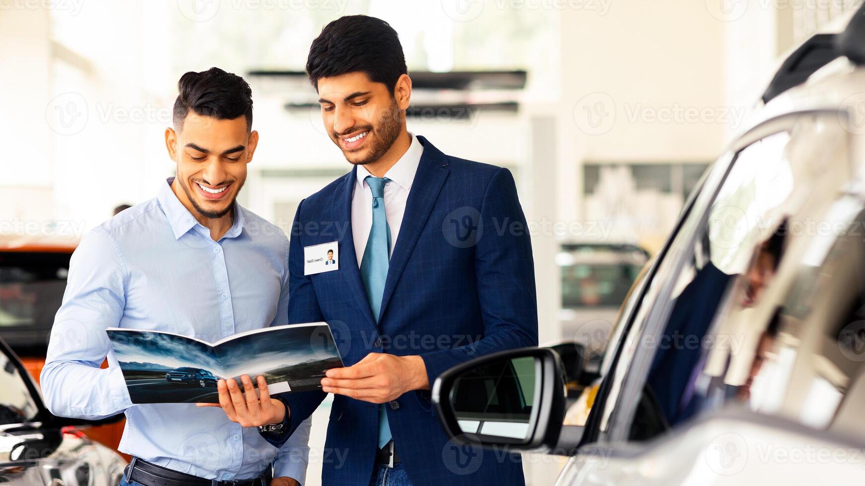 Middle-eastern guy having conversation with sales assistant in auto showroom. Handsome car dealer showing catalogue with various luxury autos, selling nice automobiles in brand new salon photo