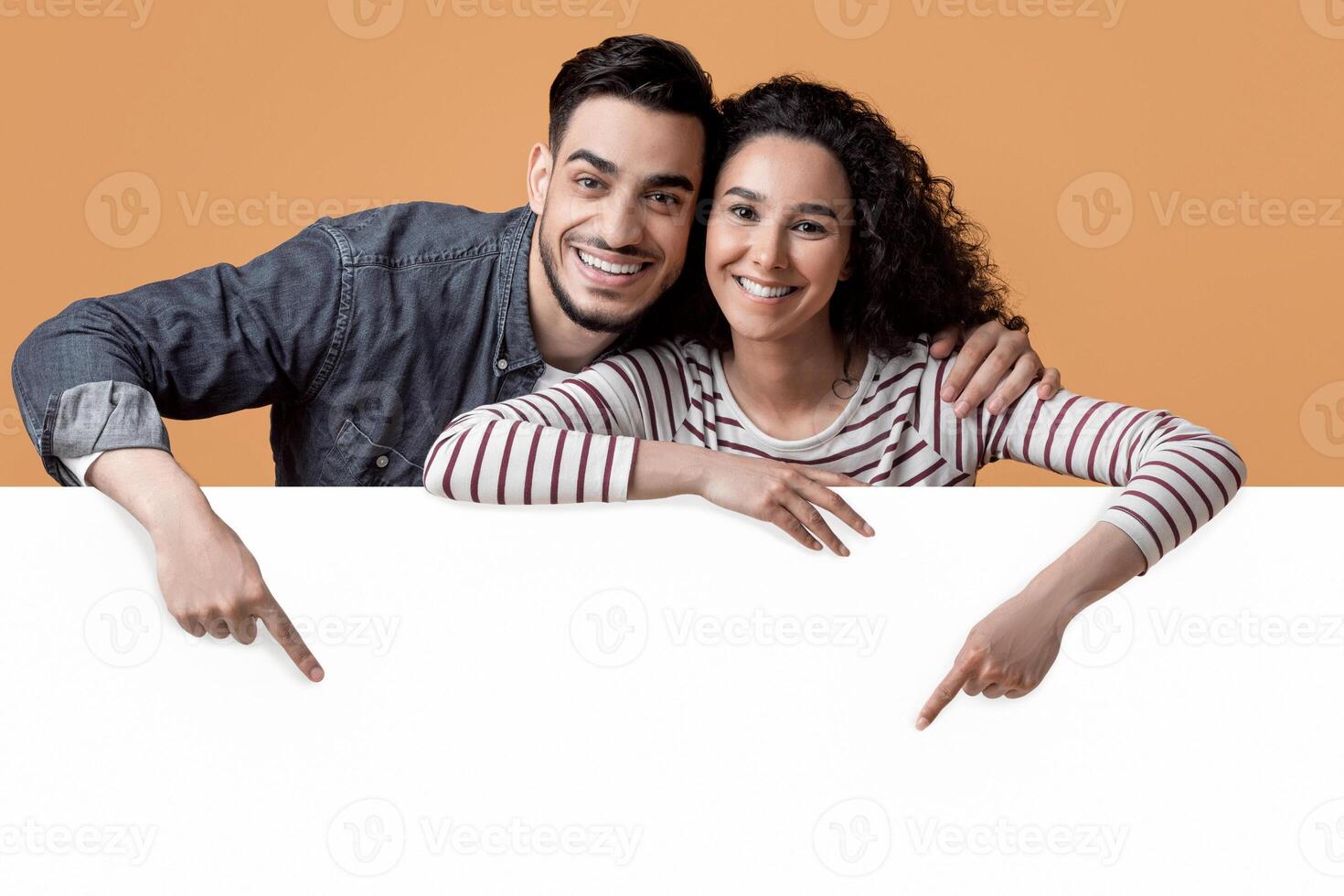 Happy Young Arab Couple Pointing Down At Big White Advertisement Board, Smiling Middle Eastern Spouses Showing Free Copy Space For Your Ad Or Design While Posing Over Yellow Studio Background photo